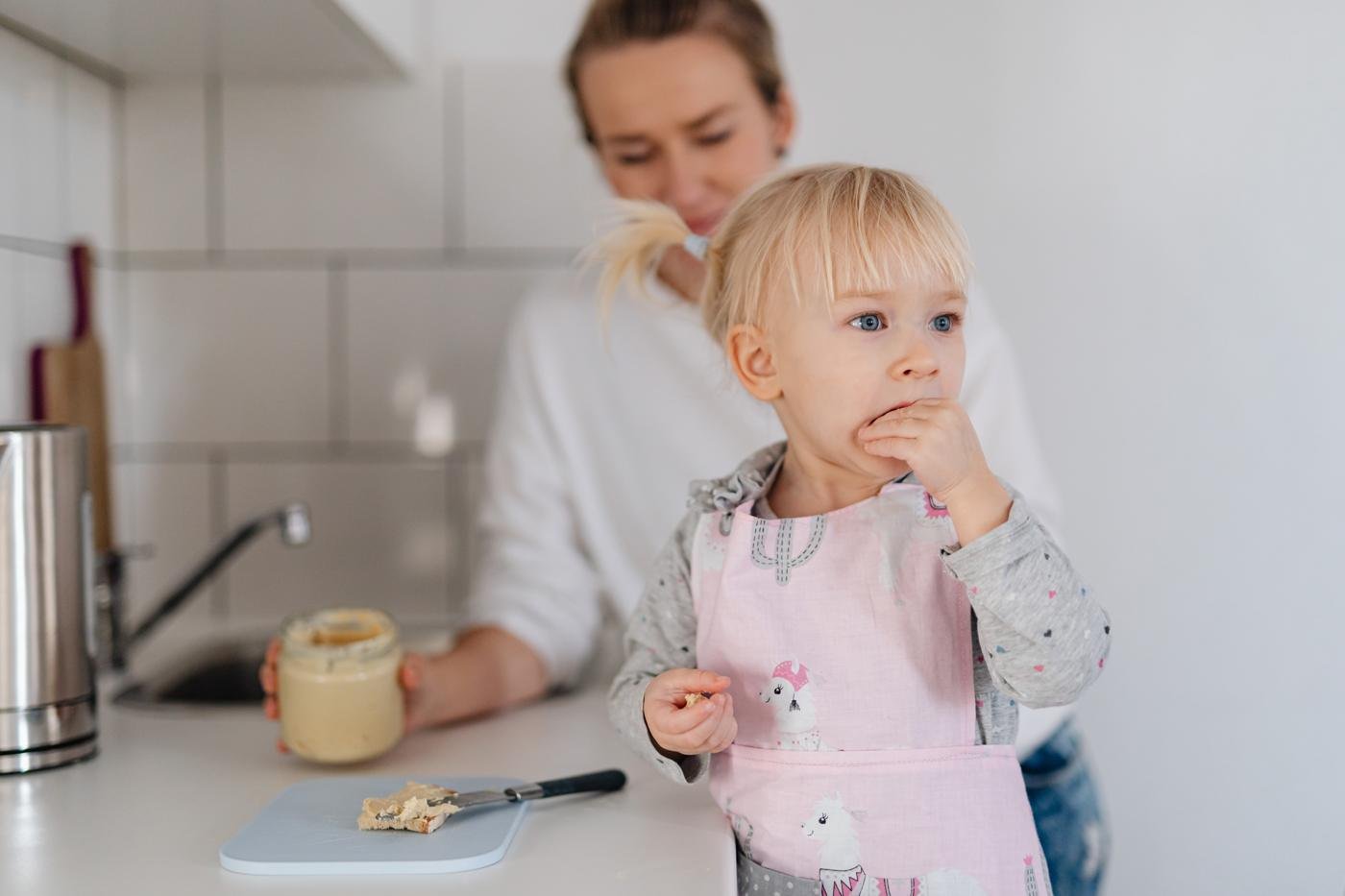 A toddler in an apron eating a snack in the kitchen while her parent looks on
